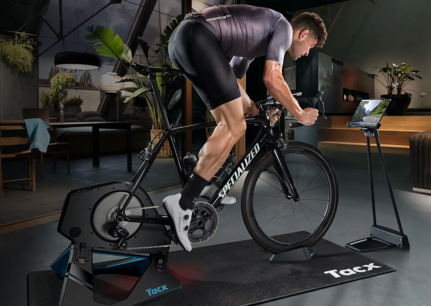 A cyclist sprinting out of the saddle on a Tacx indoor trainer beside a tablet running the training app.
